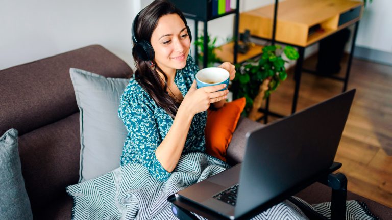 Eine Person sitzt mit Decke und Tasse auf einem Sofa. Vor ihr steht auf einem Metallständer ein Laptop.