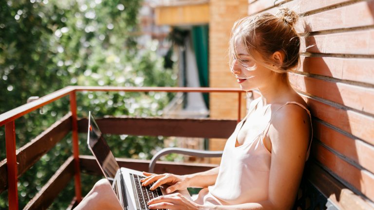 Eine Person sitzt mit Laptop auf dem Schoß auf einem Balkon. Sie wird von der Sonne angeschienen.