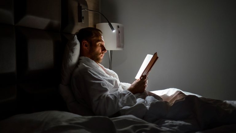 Eine Person liegt im Bett und liest. Eine Lampe, die an der Wand angebracht ist, schenkt Licht.