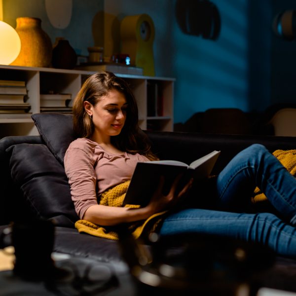 Eine Person liegt auf einer Couch und h&auml;lt ein Buch in der Hand. Hinter ihr steht eine Lampe mit warmem Licht.