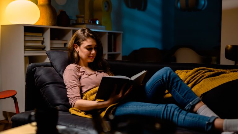 Eine Person liegt auf einer Couch und hält ein Buch in der Hand. Hinter ihr steht eine Lampe mit warmem Licht.