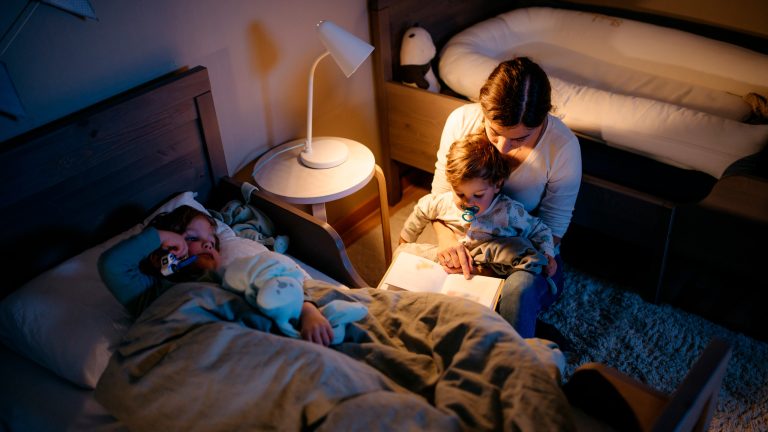 Eine Person liest zwei Kindern aus einem Buch vor. Auf dem Nachttisch steht eine wei&szlig;e Lampe.