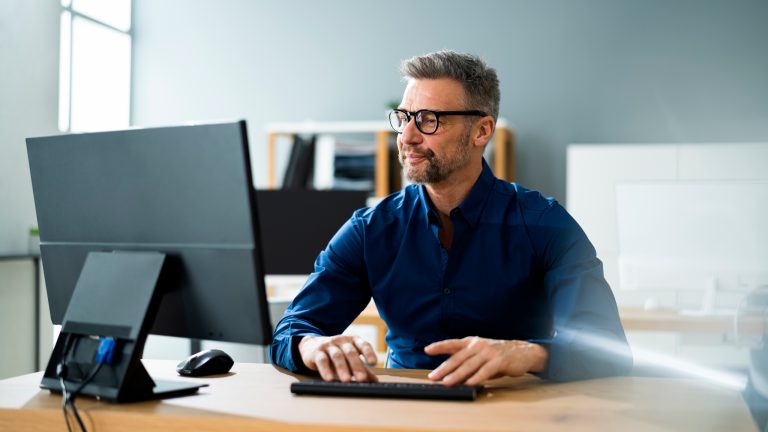 Eine Person sitzt an einem Schreibtisch, auf dem ein Monitor steht, und tippt auf einer Tastatur.