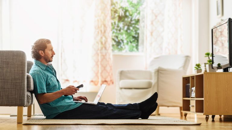 Eine Person mit einem Laptop auf dem Schoß sitzt auf dem Boden vor einem Fernseher und hält eine Fernbedienung in der Hand.