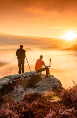 Zwei Personen schauen von einem Berg hinab in eine wolkenumzogene Landschaft und beobachten den Sonnenuntergang.