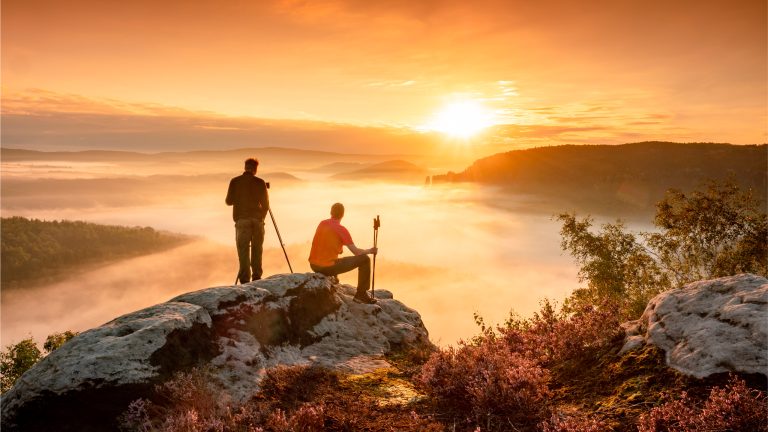 Zwei Personen schauen von einem Berg hinab in eine wolkenumzogene Landschaft und beobachten den Sonnenuntergang.