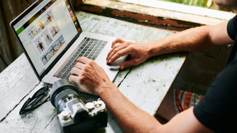 Eine Person sitzt an einem Tisch und schaut auf dem Laptop Bilder an. Daneben liegt eine Spiegelreflexkamera.