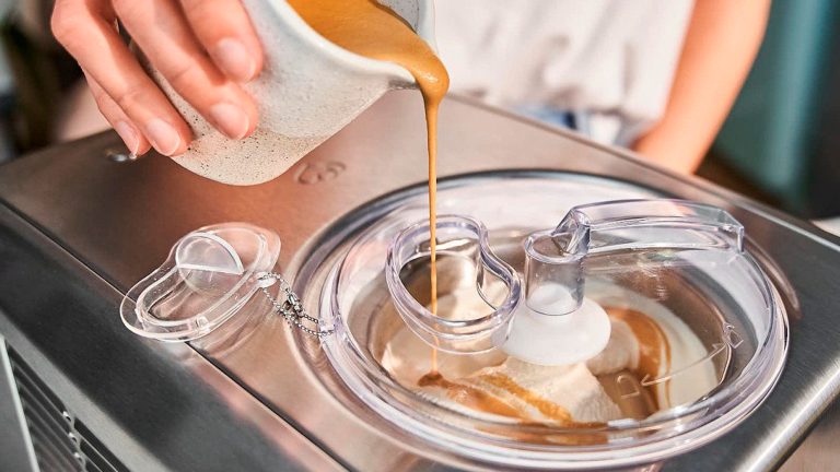 Eine Person gibt eine karamellfarbene Fl&uuml;ssigkeit in eine Eismaschine, w&auml;hrend diese die Eismasse r&uuml;hrt. 