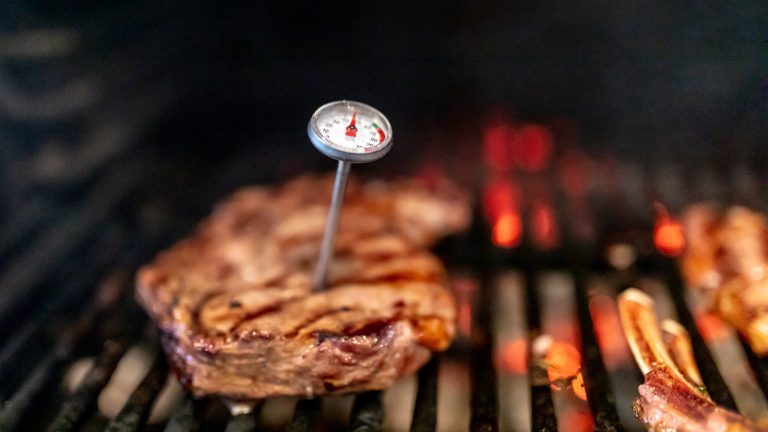 In einem St&uuml;ck Fleisch, das auf einem Grill liegt, steckt ein Fleischthermometer.