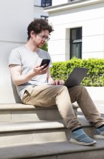 Eine Person sitzt mit einem MacBook Pro draußen und hält ihr Smartphone in der Hand.