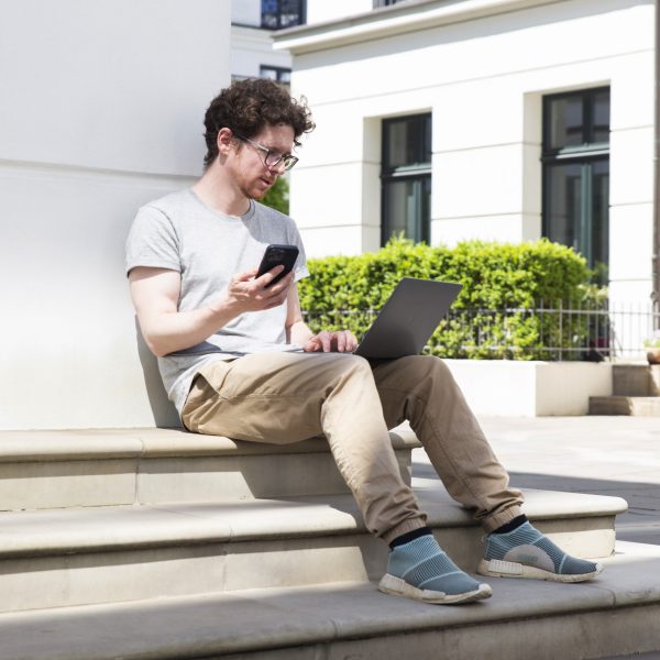 Eine Person sitzt mit einem MacBook Pro drau&szlig;en und h&auml;lt ihr Smartphone in der Hand.