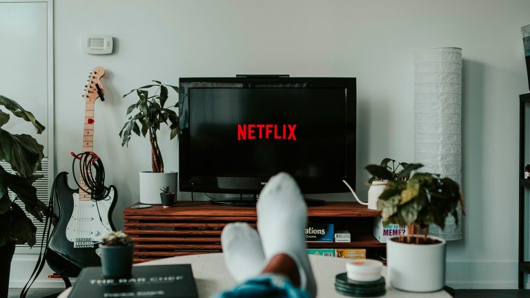 Blick auf einen Fernseher, auf dem Netflix läuft. Im Vordergrund sind die Füße einer Person auf einem Tisch zu sehen.