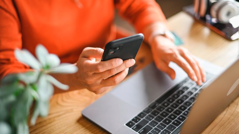 Eine Person hält ein iPhone in der Hand und bedient mit der anderen ein MacBook Pro.