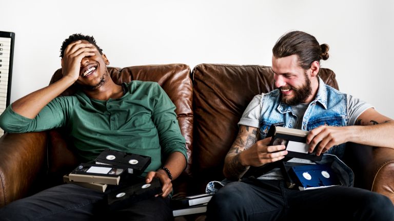 Zwei Personen sitzen auf einem Sofa und halten einige VHS-Kassetten in den H&auml;nden.