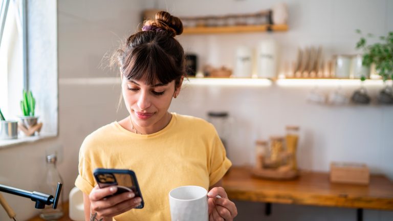 Eine Person steht mit einer Tasse in der Hand in einer Küche und blickt auf ein Smartphone, das sie in der anderen Hand hält.