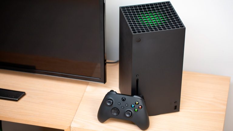 Eine Xbox Series X steht neben einem Fernseher auf einem Bücherregal. Ein Controller lehnt an der Konsole.