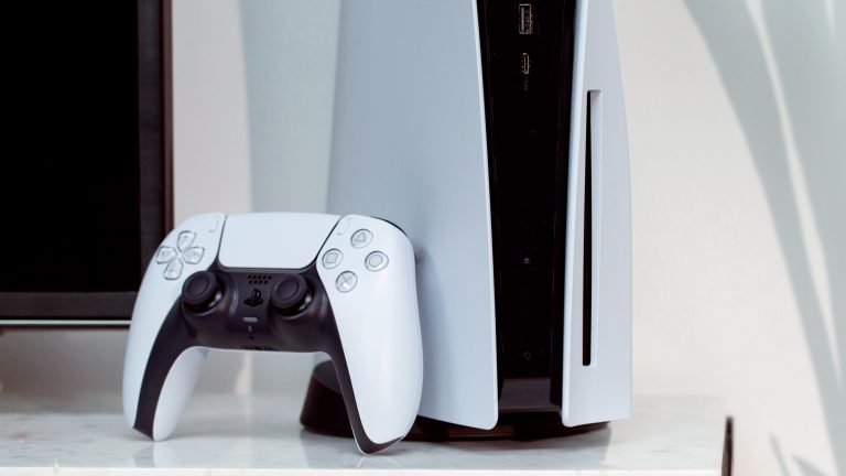 Eine PlayStation 5 steht auf einem Marmortisch neben einem Fernseher. An der Konsole lehnt ein DualSense-Controller.