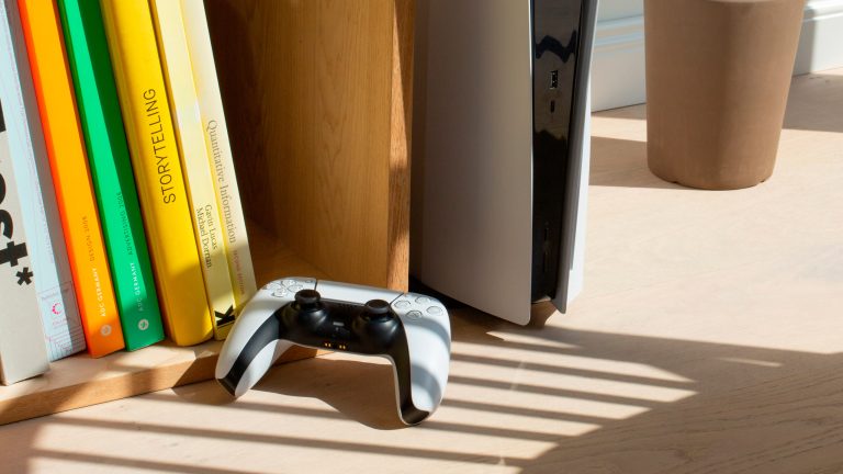 Neben einem Bücherregal ist eine PS5 aufgestellt. Davor sonnt sich im Halbschatten ein DualSense-Controller.