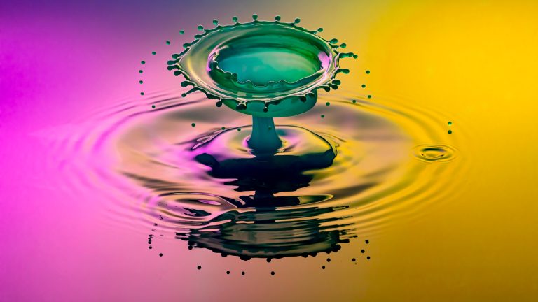 Ein Wassertropfen erzeugt einen Planscher in magenta-gelb beleuchtetem Wasser.