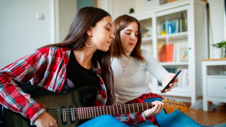 Zwei Personen sitzen auf dem Boden. Eine h&auml;lt eine E-Gitarre in der Hand, die andere ein Smartphone hoch, auf dem wohl Noten zu sehen sind.