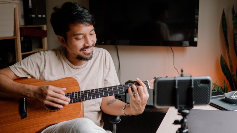 Eine Person sitzt mit einer Gitarre in der Hand vor einem Smartphone.