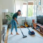 Eine Person saugt einen kleinen Wohnraum, auf dessen Boden ein grauer Teppich liegt.
