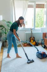Eine Person saugt einen kleinen Wohnraum, auf dessen Boden ein grauer Teppich liegt.