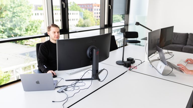 In einem Büro sitzt eine Person vor einem Ultrawide-Monitor. Schräg gegenüber sitzt eine zweite Person vor zwei Monitoren.
