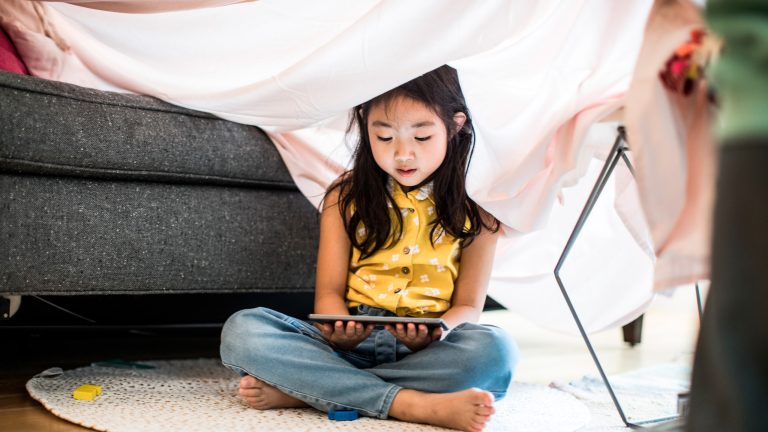 Ein Kind sitzt unter einer Decke, die über ein Sofa gespannt ist und schaut sich etwas auf einem Tablet an.