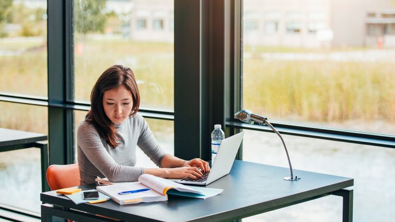 Eine Person sitzt in einer Universit&auml;t, schaut in B&uuml;cher und bedient einen Laptop.