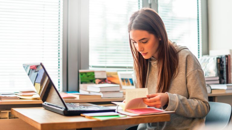Eine Person sitzt mitsamt einigen Notizbl&ouml;cken und B&uuml;chern vor einem Laptop.