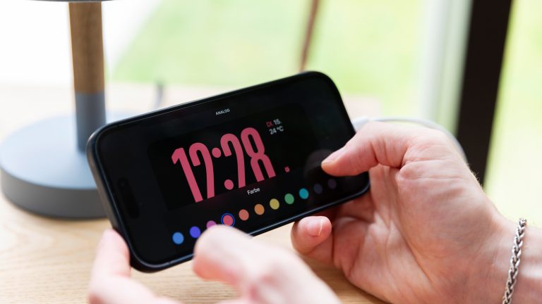 Eine Person hält ein iPhone 14 Pro in den Händen, auf dem gerade die Farbe einer Uhrdarstellung im Standby-Modus geändert wird.