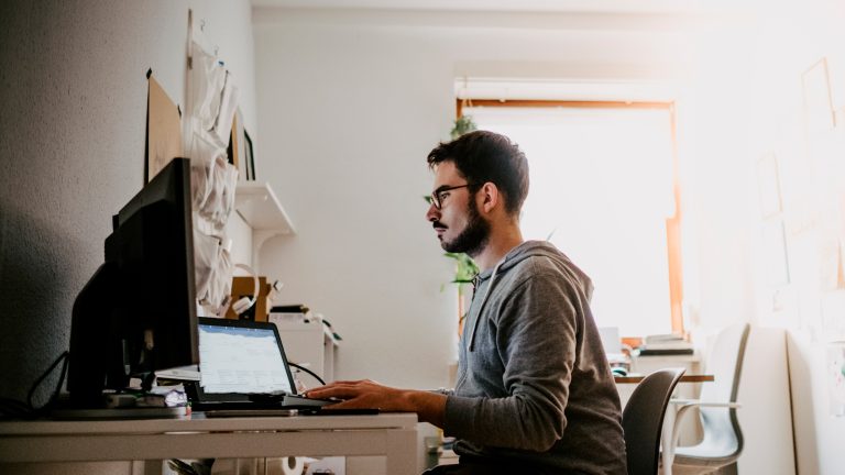 Eine Person sitzt vor einem Schreibtisch, auf dem ein Laptop und ein Monitor zu sehen sind.