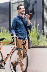Eine Person führt mit einer Hand ein Fahrrad mit sich, in der anderen hält sie ein Smartphone, in das sie spricht.