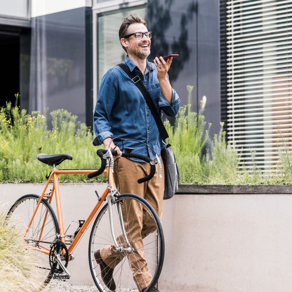 Eine Person f&uuml;hrt mit einer Hand ein Fahrrad mit sich, in der anderen h&auml;lt sie ein Smartphone, in das sie spricht.