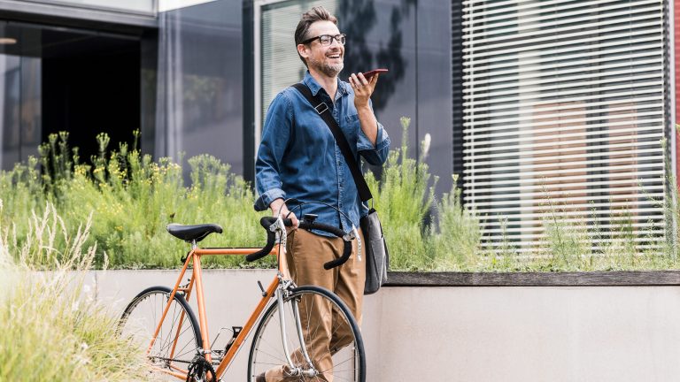 Eine Person führt mit einer Hand ein Fahrrad mit sich, in der anderen hält sie ein Smartphone, in das sie spricht.
