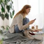 Eine Person sitzt auf einem Bodenkissen und bedient Laptop und Smartphone gleichzeitig.