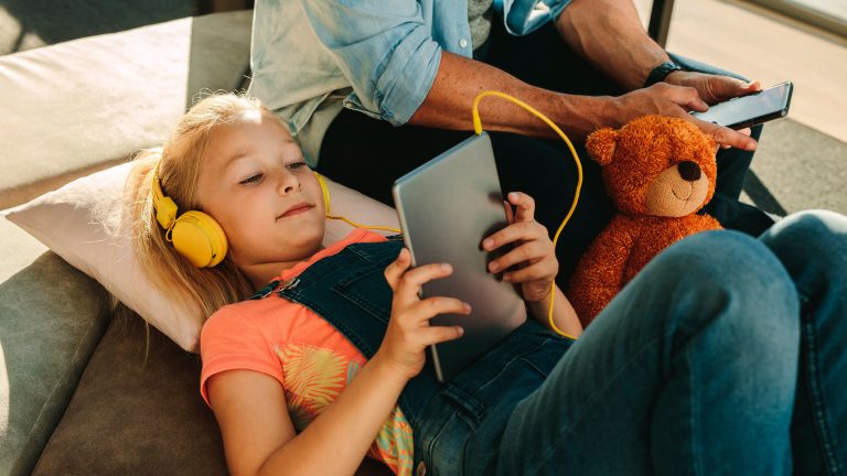 Ein Kind liegt auf einem Sofa und schaut mit Kopfhörern auf dem Kopf etwas auf einem Tablet.