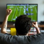 Eine Person sitzt vor einem Fernseher und jubelt. Auf dem Bild ist ein Football-Spiel zu sehen.