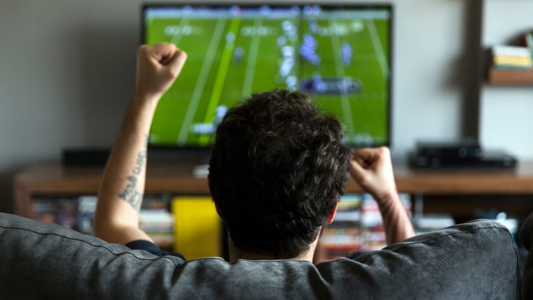 Eine Person sitzt vor einem Fernseher und jubelt. Auf dem Bild ist ein Football-Spiel zu sehen.