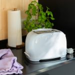 Ein wei&szlig;er Toaster steht neben einer K&uuml;chenrolle und einem lilafarbenen Putztuch auf einer schwarzen K&uuml;chenanrichte.
