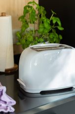 Ein weißer Toaster steht neben einer Küchenrolle und einem lilafarbenen Putztuch auf einer schwarzen Küchenanrichte.