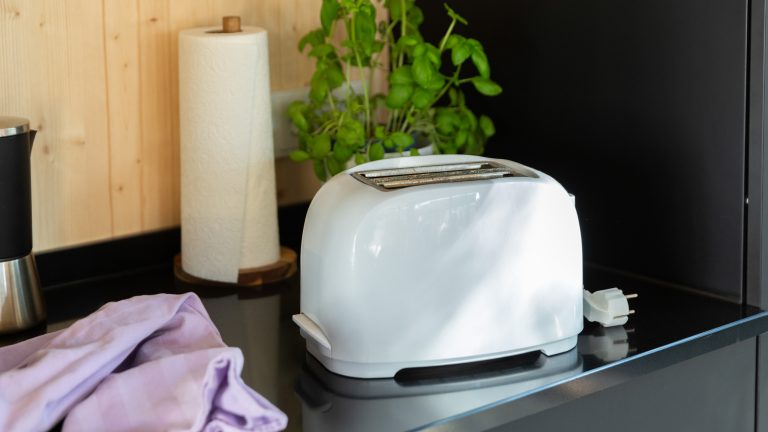 Ein weißer Toaster steht neben einer Küchenrolle und einem lilafarbenen Putztuch auf einer schwarzen Küchenanrichte.