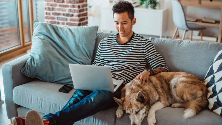 Eine Person sitzt zusammen mit einem Hund auf einem Sofa und schaut in einen Laptop.