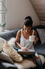 Eine Person mit einem Kaffee in der Hand sitzt auf einem Sofa und bedient einen Laptop.