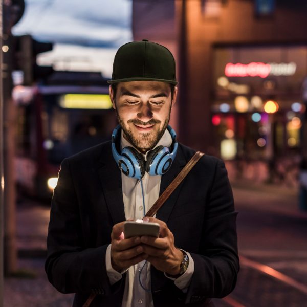 Eine Person mit Kopfhörern um den Hals steht vor öffentlichen Verkehrsmitteln und schaut auf ihr Smartphone.