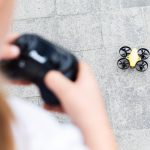 Ein Kind hält eine Fernbedienung in der Hand. Die gehört zu einer Drohne, die vor ihm auf dem Boden steht.