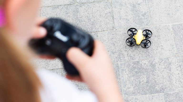 Ein Kind hält eine Fernbedienung in der Hand. Die gehört zu einer Drohne, die vor ihm auf dem Boden steht.