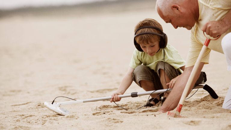 Ein Kind und eine ältere Person buddeln im Sand an einem Strand. Das Kind hält einen Metalldetektor in den Händen.