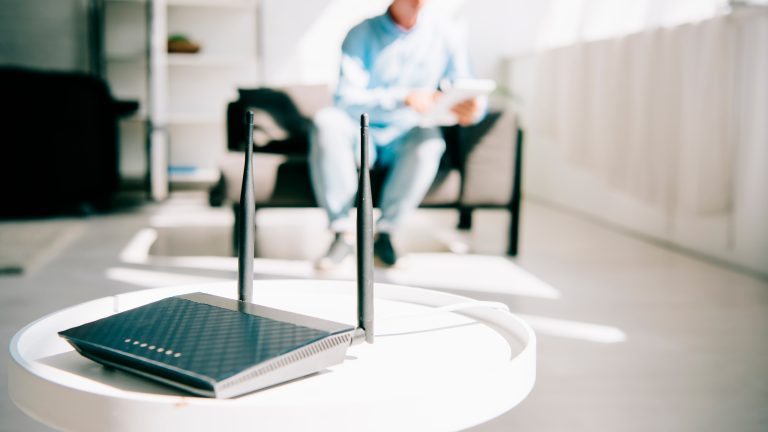 Ein Router steht auf einem kleinen Tisch. Im Hintergrund ist eine Person an einem Tablet zu sehen.